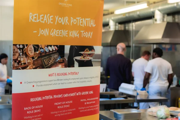 A display banner for the Greene King "Release Your Potential" program, which offers employment in the hospitality industry to ex-offenders, set against a background of staff working in a commercial kitchen.