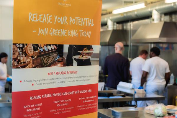 A display banner for the Greene King "Release Your Potential" program, which offers employment in the hospitality industry to ex-offenders, set against a background of staff working in a commercial kitchen.