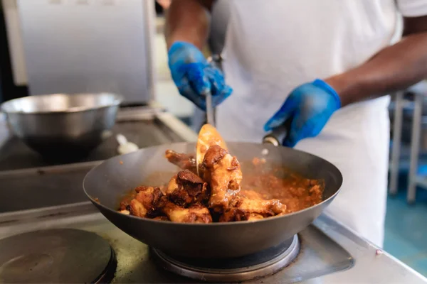 Person cooking chicken in a pan on a stovetop, stirring with gloved hands.