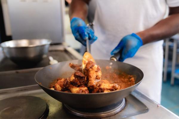 Person cooking chicken in a pan on a stovetop, stirring with gloved hands.