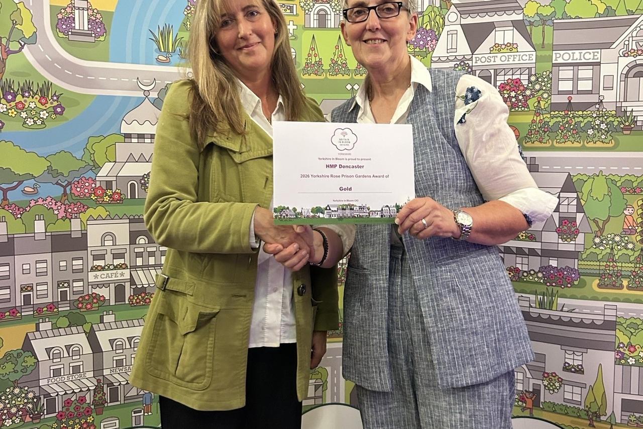 A Novus and Serco colleague stood together holding the Yorkshire in Bloom Gold Award, whilst stood in front of a Yorkshire in Bloom banner