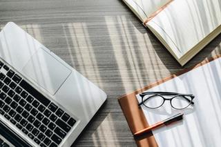An open book, paper, glasses and pen placed on a table alongside a grey laptop
