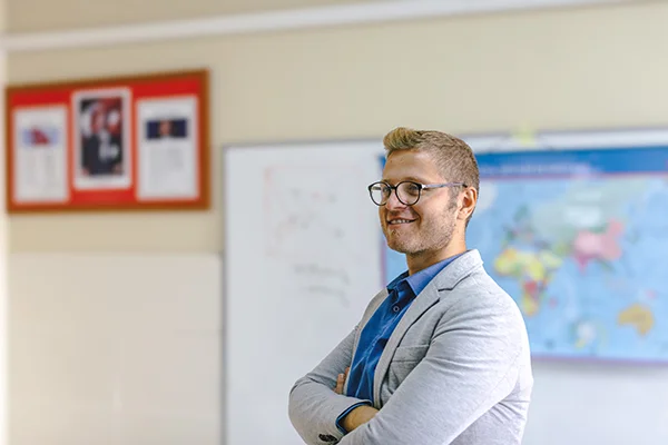 Happy young male teacher standing behind a whiteboard in a school classroom, symbolising engaging and supportive teaching in education.