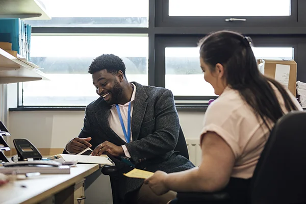 Two people collaborating in an admin office, with one focused on paperwork and the other talking.