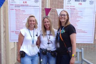 3 colleagues stood together outside, smiling at the camera with the HMP Stafford Soundwave event posters in the background posted on the fencing behind them