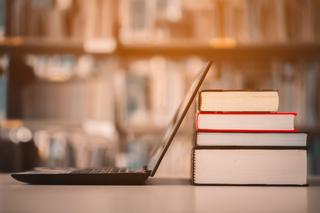 A laptop with its screen leaning against a pile of books on a table.