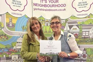 A Novus and Serco colleague stood together holding the Yorkshire in Bloom Gold Award, whilst stood in front of a Yorkshire in Bloom banner