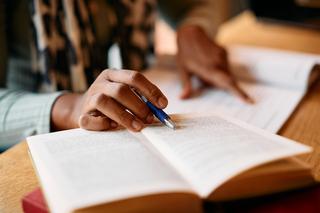 An individual working in a notebook and reading a book with a pen following each word