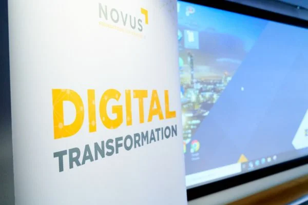 Sign that reads "Novus Digital Transformation", situated next to a big screen