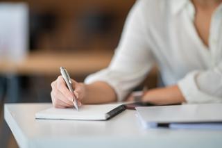Woman in white shirt, sat at a desk writing in a notebook.
