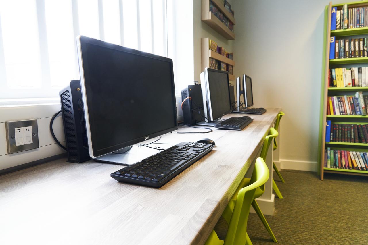 A row of desktop computers and keyboards positioned on a long workstation in a learning or library space, with bright green chairs and bookshelves filled with books in the background.