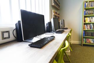 A row of desktop computers and keyboards positioned on a long workstation in a learning or library space, with bright green chairs and bookshelves filled with books in the background.