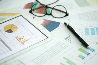 Papers and a tablet scattered across a table which consist of data including pie charts and bar charts whilst a pair of glasses and a pen sit on top of some of the papers