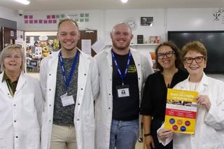 A group of five staff members stand together in a classroom at HMP Brinsford. Three people are wearing white chef coats, with one person holding a yellow promotional poster.