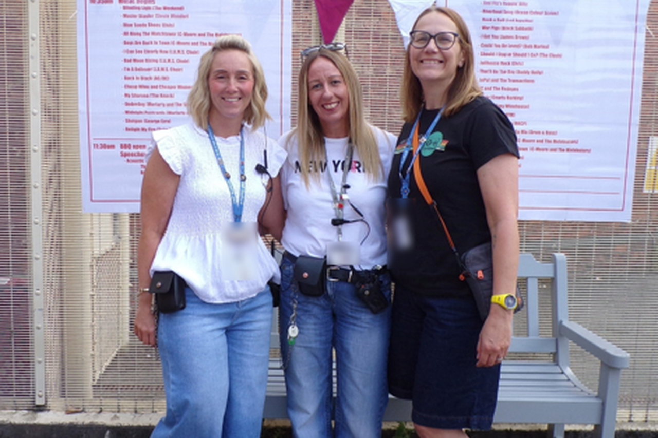 3 colleagues stood together outside, smiling at the camera with the HMP Stafford Soundwave event posters in the background posted on the fencing behind them