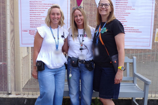 3 colleagues stood together outside, smiling at the camera with the HMP Stafford Soundwave event posters in the background posted on the fencing behind them