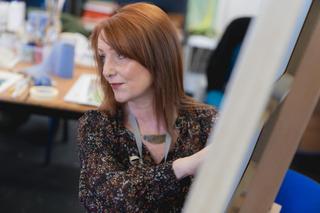 Staff member guiding a learner through a creative enrichment activity beside an easel in a classroom at HMP Styal.