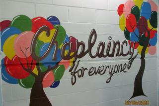 White wall with a colourful mural covering it with the phrase 'Chaplaincy for everyone' painted in brown and two trees painted on either side with multi-coloured branches
