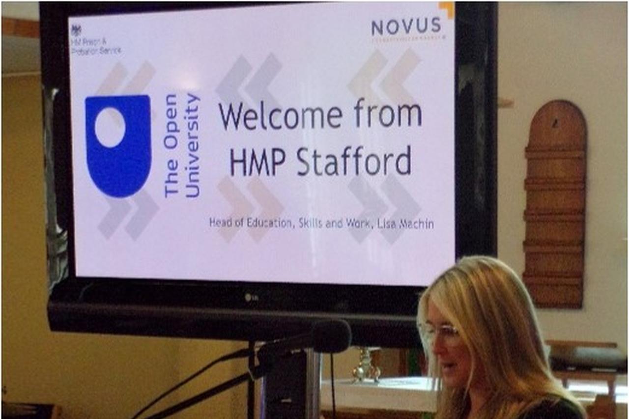 Novus colleague stood behind a podium presenting the HMP Stafford Graduation with a screen in the background with the words 'Welcome from HMP Stafford'