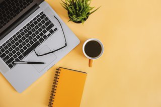 A computer with glasses and a pen lay on top of it, a mug, a notebook and a plant all positioned together on top of a yellow background