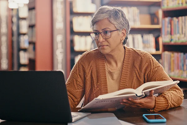 Woman in a library holding a book and working on a laptop, representing the 'Great Pensions' benefit of working for Novus in prison education.