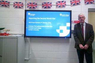 A history lecturer stood in a classroom, in front of a presentation about reporting in the Second World War