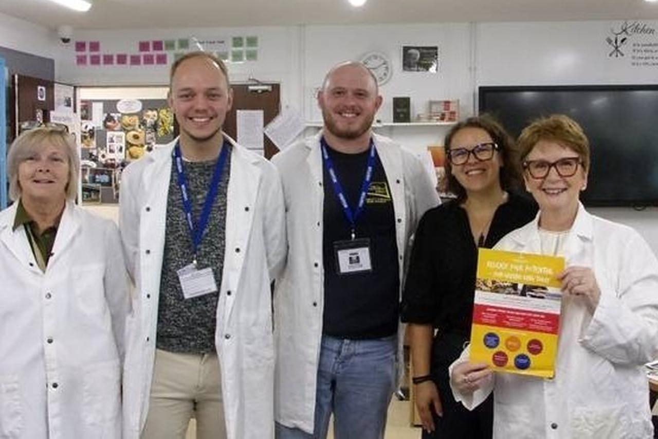 A group of five staff members stand together in a classroom at HMP Brinsford. Three people are wearing white chef coats, with one person holding a yellow promotional poster.