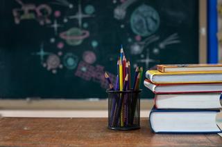 A pile of books and a pencil holder full of colourful pencils sat on a table with a blackboard behind covered in drawings of the different planets, earth and stars