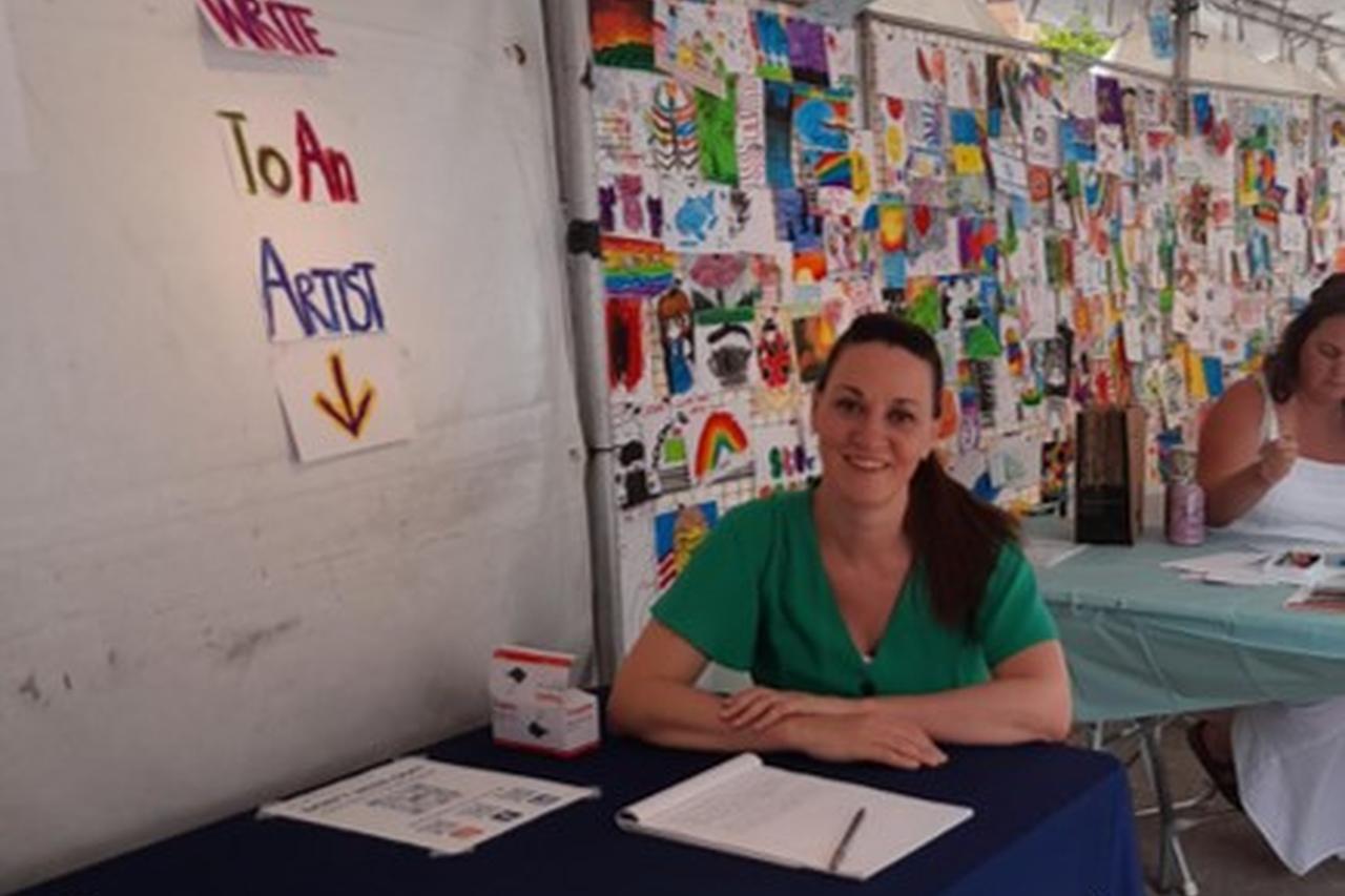 Sarah Hartley sat at a table in a tent with a wall of artwork behind her at the Ann Arbor Summer Fair in Michigan USA